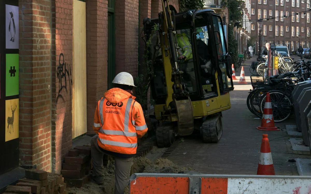 Un muncitor la terasamente și un operator cu un miniexcavator lucrând pe o stradă